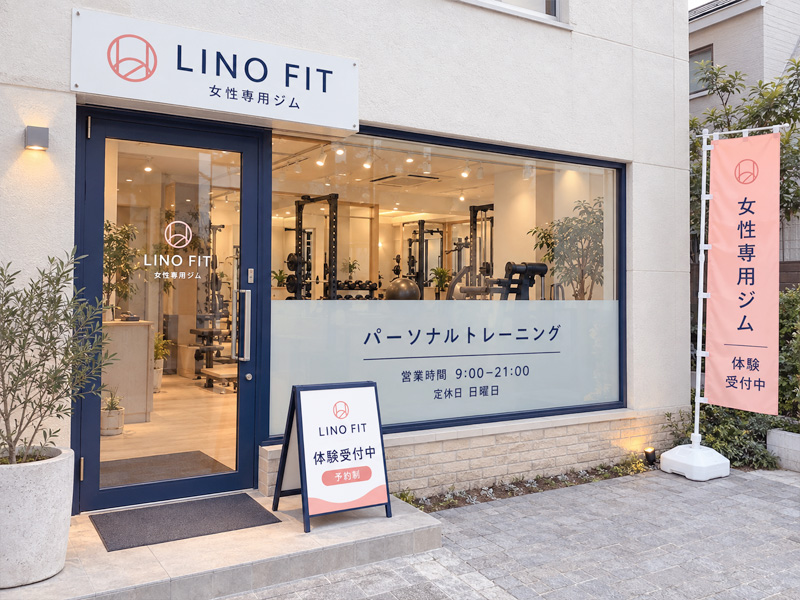 白い外壁とネイビーの建具に、プレート看板・ウィンドウサイン・体験案内のスタンド看板・のぼり旗を組み合わせた女性専用ジムLINO FIT